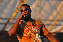 Gucci Mane holds a microphone onstage. He is wearing his ice cream cone pendant necklace but does not yet have the tattoo on his face.