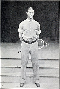 A man in white pants and shirt swings a wooden racket on a grass court, with other male and female tennis players in the background