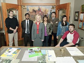 A group of people stand in an office-style room in front of a table with documents and booklets. From left to right stand a man with arms crossed wearing a black shirt, a man wearing a gray suit and green tie with his arms behind his back, a woman in a gray suitcoat and red shirt, a woman with a gray jacket and purple shirt, a man with arms crossed wearing a tan suit, a young girl wearing a blue blouse and gray shirt and a seated man wearing a white T-shirt with red sleeves.