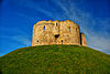 A tall, circular, roofless building of honey coloured stone positioned on top of a high mound of grass.