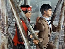 A man dressed in a scarlet uniform and cap holds a suited male civilian hostage, using the civilian as a shield while pointing a gun at an off-screen enemy. The agent and hostage are standing on a metal structure, which is shown to be high above the ground due to the scale of the buildings and streets below them.