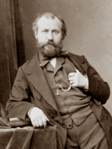 Man in early middle age, slightly balding, with neat moustache and beard, standing facing the viewer