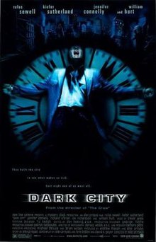 A black poster. Above reads the lines: "Rufus Sewell", "Kiefer Sutherland", "Jennifer Connelly", "and William Hurt". In the center, against a black background, a man wearing a blue jacket is rested against an upright clock with Roman numerals as big as him; the setup cast in a blue tint. His arms are outspread, and his head is tilted back with his mouth agape. Behind the man and the clock is a dark city skyline. Below them is the tagline, "They built the city to see what makes us tick. Last night one of us went off." Below the tagline is the film title, "Dark City".