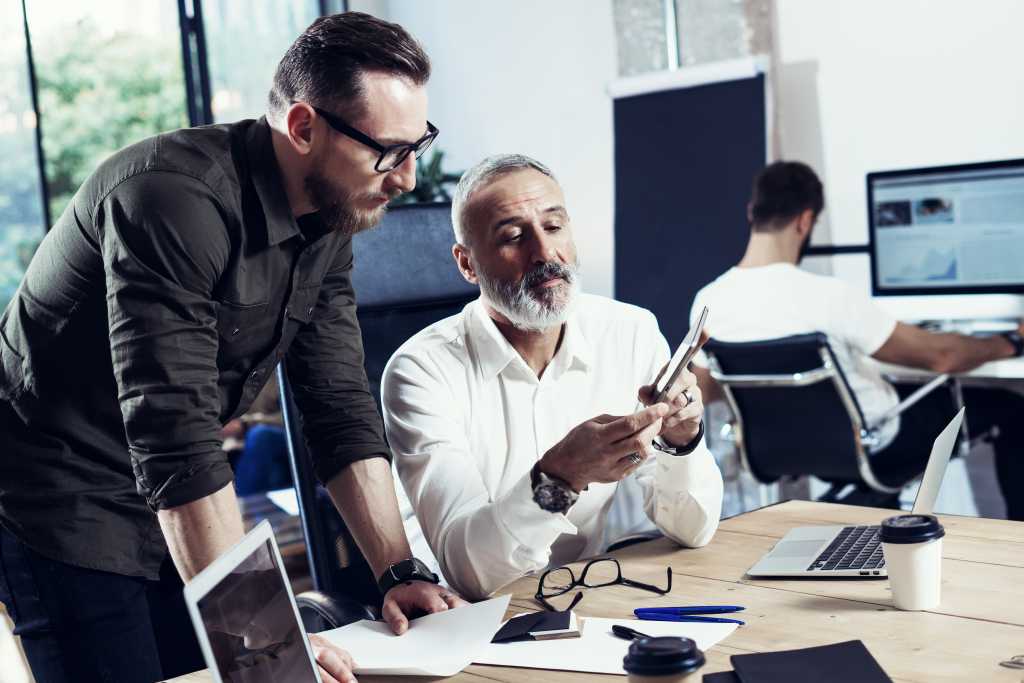 Group of two coworkers making great time brake during work process in modern office.Adult bearded man using mobile tablet with young partner.Business people meeting process.Horizontal, blurred
