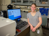 Grace Waters poses smiling in the lab, leaning against a desk with a computer monitor showing data and graphs. 