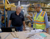 Two men stand a table in a warehouse space, looking at a broken piece of concrete that one is holding. 