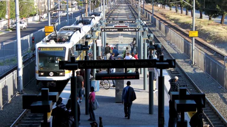 An overhead view of a Blue Line light rail station in Los Angeles, with people walking on the platform boarding and disembarking a train in the left-hand side of the frame