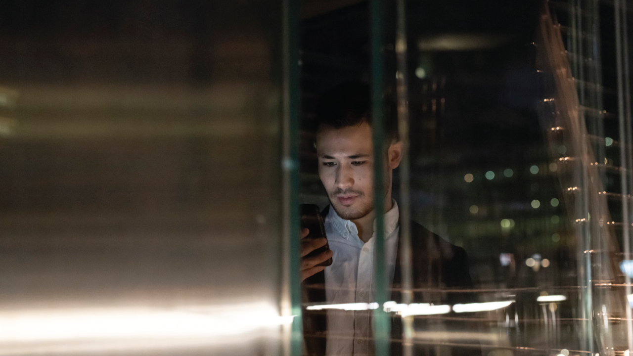 A photo of a young businessman in office building at night looking at smartphone, window view, Milan, Italy.