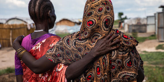 Two women stand in a open space with their backs to the camera. They have an arm around each other's back.