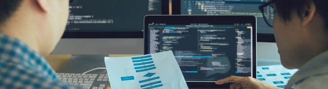 Two engineers examining a process flow on paper with coding on computer screens in the background.