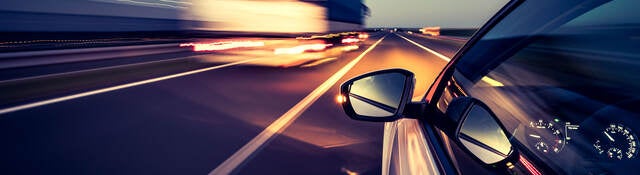 Silver car with side-view mirror driving on highway beside heavy duty truck at night