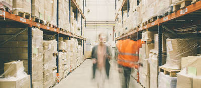 Two people walking through a warehouse of packages on shelves