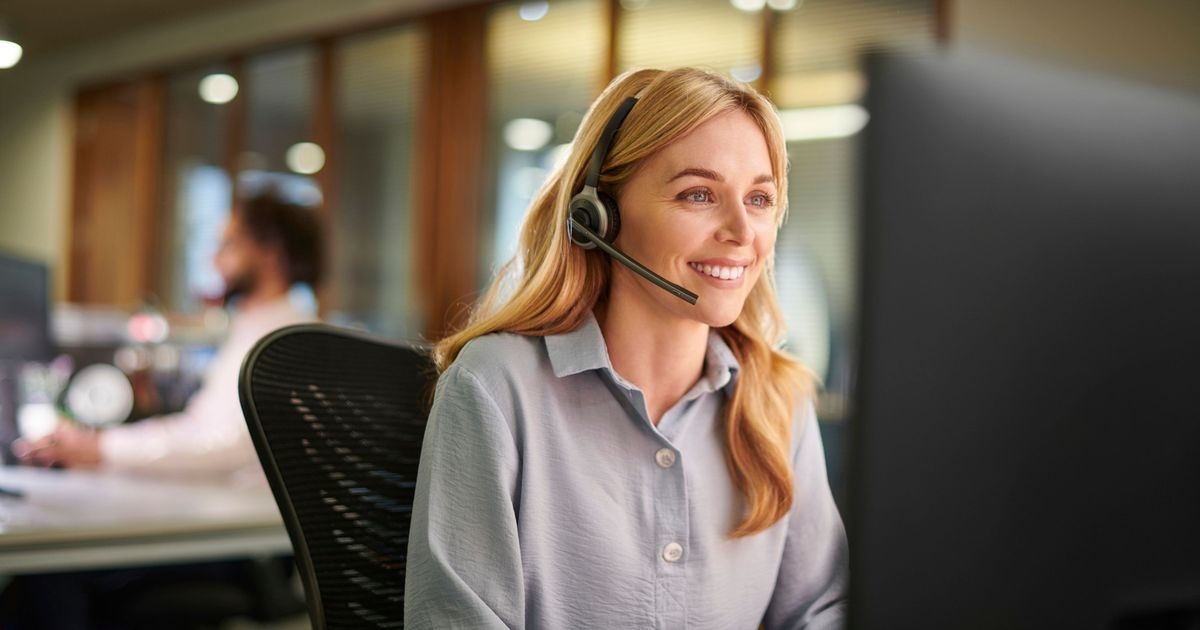 Eine Frau mit Headset sitzt lächelnd an einem Arbeitsplatz, im Hintergrund ein weiterer Mitarbeiter.