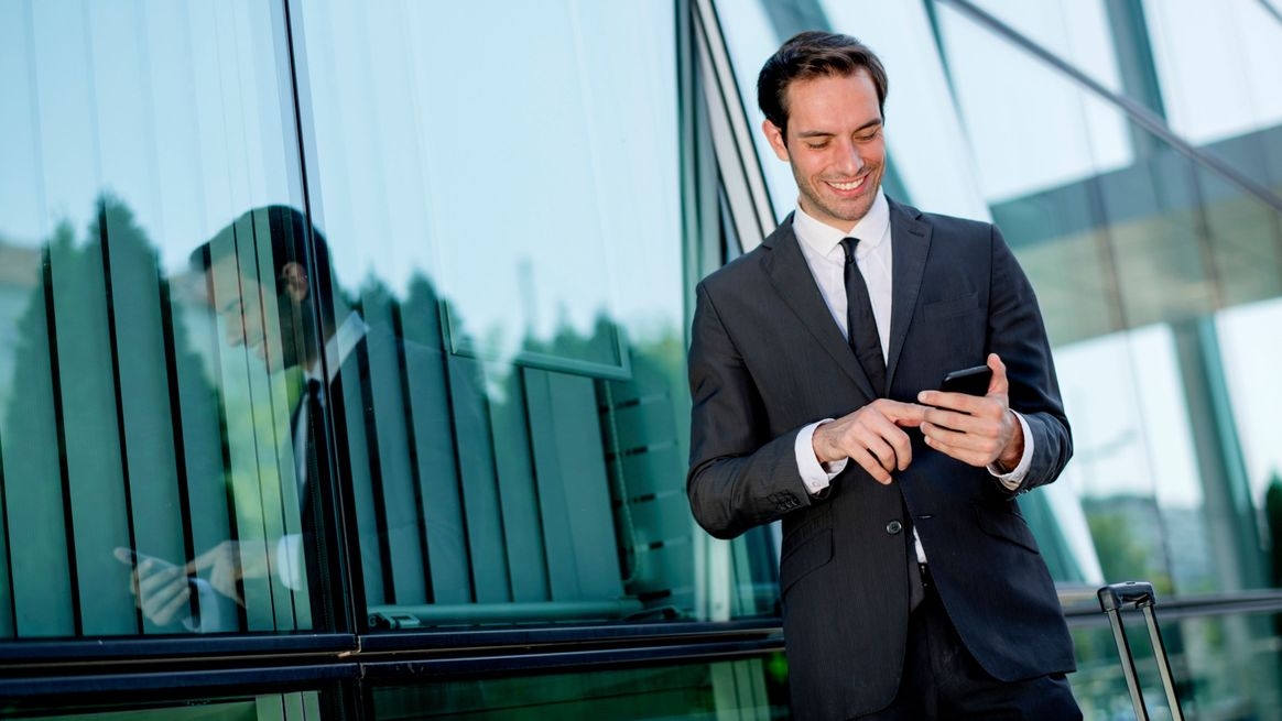 Ein Geschäftsmann steht mit einem Smartphone in der Hand vor einer Glaswand und tippt etwas