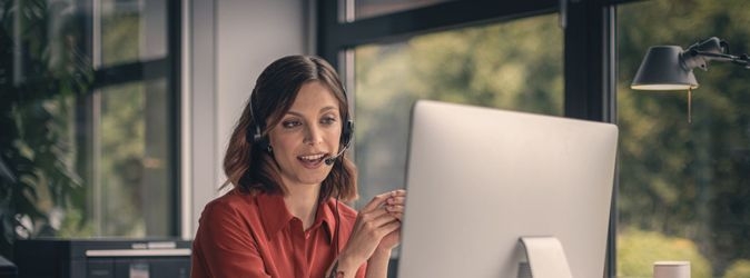 Eine Frau in roter Bluse sitzt an einem Mac und telefoniert via Headset 