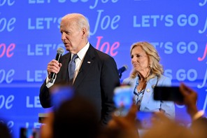 Joe Biden wearing a suit and speaking into a microphone in front of a backdrop reading "Let's Go Joe"; Jill is seated behind him.