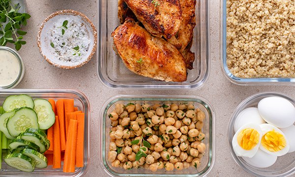 Glass dishes in two rows containing food that would constitute nutritious meals nurses to sustain nurses at work. There is thyme and yoghurt dress, chicken breast, couscous, cucumber, carrot, chickpeas and boiled eggs