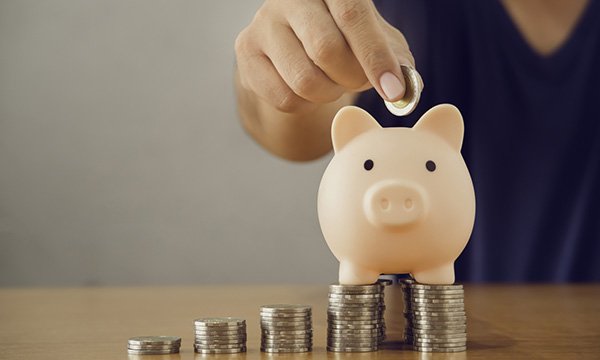 A picture of a pig-shaped money box sitting on top of coin stacks of decreasing size, with a person’s hand hovering above about to place a £2 coin inside the money box