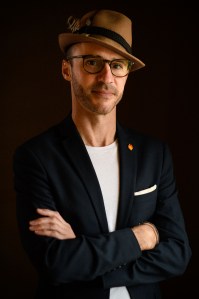 Headshot photograph of John Mills, co-founder and CEO of Watch Duty. He has is arms crossed and is wearing dark framed glasses, a jaunty straw hat, and a dark blazer over a white t-shirt.