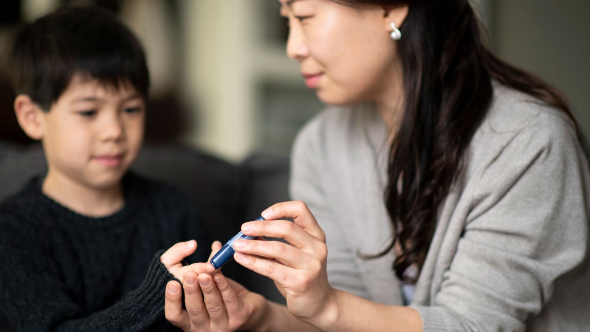 A mother and son are sitting together in a living room. She is helping him check his blood sugar levels because he is diabetic.