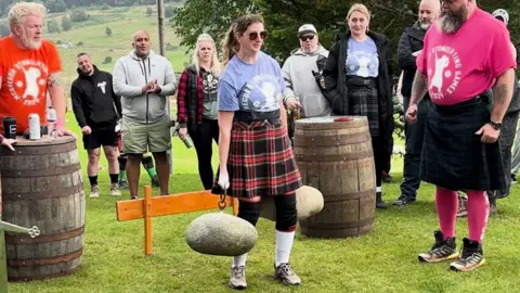 Gemma Doherty, wearing a kilt and blue t-shirt lifts two large stones on chains.