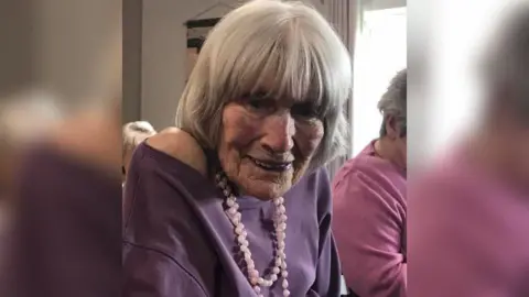 Hilda Bond smiling at the camera, she appears to be in the care home as there are other elderly residents around her. She is wearing a purple jumper with a pink beaded necklace around her neck. Her hair has been styled and lays flat and she has a fringe