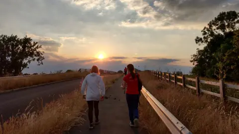 Georgeta Butland MONDAY - Two women walk away from the camera, one is in a long sleeve white top, the other is in a red t-shirt. They both wear black tracksuit bottoms and trainers. The sun is setting on the horizon over a motorway bridge. To the left of the picture is a road and on either side the verges are dry yellowing long grass.
