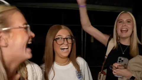 Three women celebrate England's Euro 2024 win, one has her arm in the air.