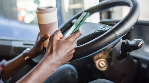 A man sits in a stationary vehicle in front of a steering wheel holding a cup of coffee in one hand and a phone in the other