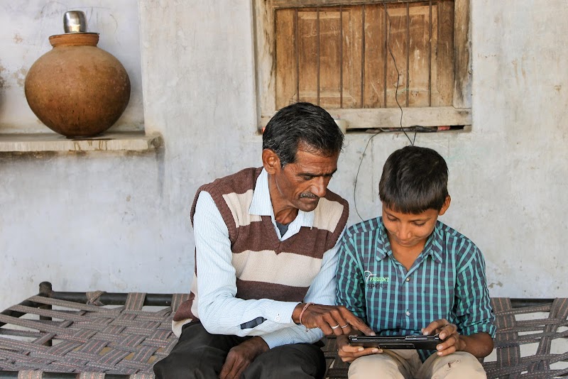 Young boy in plaid shirt looking at tablet and getting assistance from man in striped vest seated to his right.