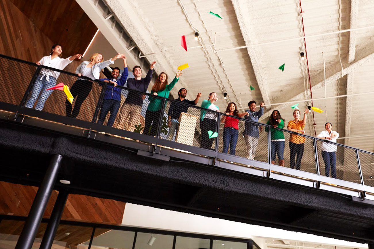 A group of Googlers stand at the edge of an indoor balcony. They toss paper airplanes over the balcony.