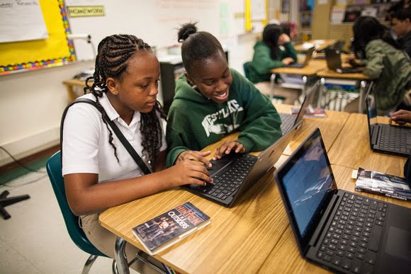Hai nữ sinh cấp hai đang dùng chung máy tính xách tay trong một lớp học. Cô gái mặc áo polo trắng đang tập trung vào màn hình. Bạn của cô mặc chiếc áo hoodie màu xanh lá cây đang mỉm cười trước màn hình.