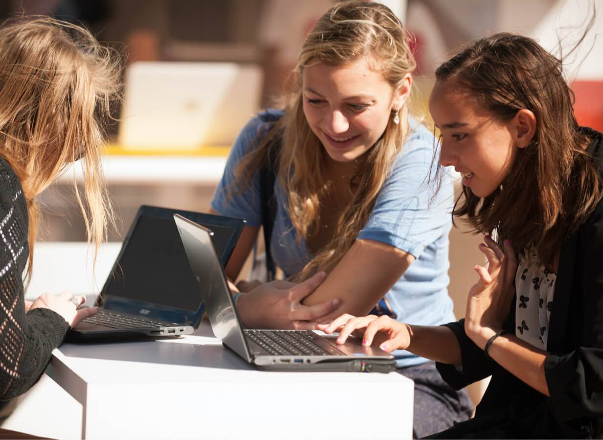 Três alunas em uma mesa ao ar livre trabalhando em Chromebooks.