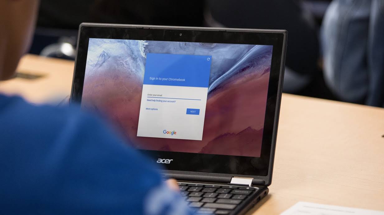 Plano fechado de um aluno acessando a tela de login do Google em um Chromebook sobre a mesa.