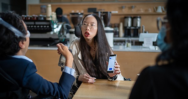 A woman with long brown hair and glasses sits at a table in a coffee shop. She’s holding a phone in her left hand with the screen facing the two people sitting in front of her.