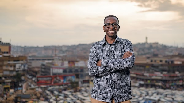 Инженер Байномугиша стоит со скрещенными на груди руками и улыбается в камеру. Позади него видны очертания города Кампала в Уганде.
