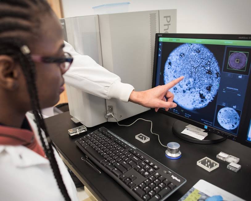 Dedo apuntando a la pantalla de un ordenador que una mujer mira de cerca. Parece que están en un laboratorio científico y que observan el primer plano de una masa circular.