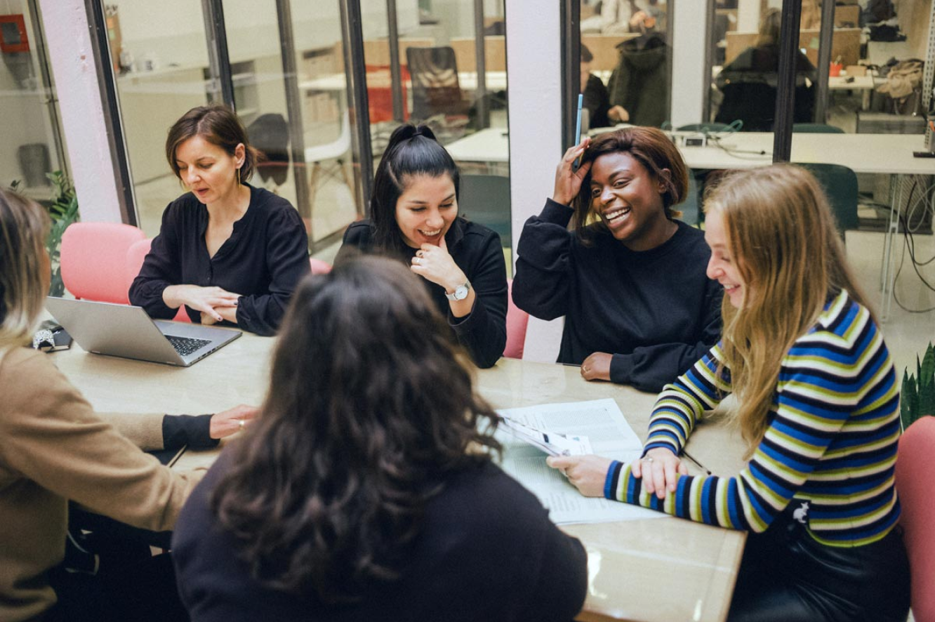 Un grupo de mujeres sentadas en una mesa mientras trabajan en sus laptops, sonriendo y riendo.