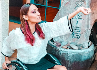 A photo of Angela Rockwood, a glamorous young woman with dyed red hair, sitting in a wheelchair holding her wrist out.
