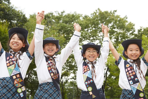 Cuatro niñas scouts japonesas cogidas de la mano con los brazos en alto en señal de celebración.