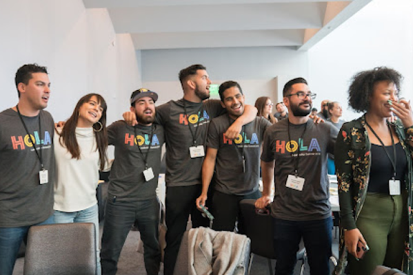 Cinco empleados de Google cantan de pie abrazados mientras otros dos Googlers están a su lado sonriendo