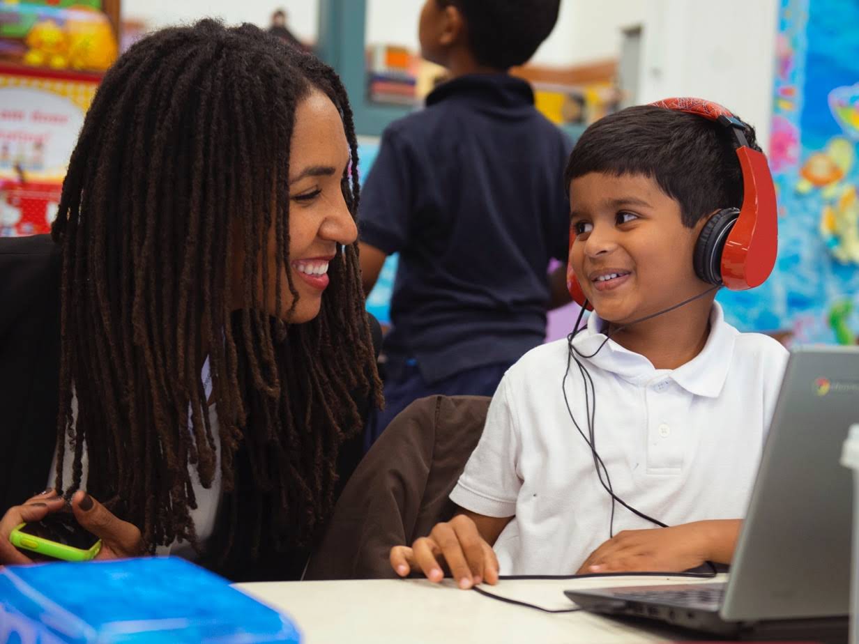 Chromebook'ta kulaklıkla bir şeyler dinlemekte olan öğrenciye gülümseyerek uzanan bir kadın.