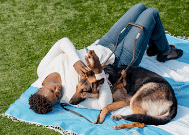 黒人女性が芝生にブランケットを敷いて介助犬のジャーマン シェパードと一緒に横たわっている。女性は Android デバイスを持っている。