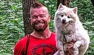 A photo of Rusty, who is a strong young man wearing a red t-shirt, holding his fluffy white dog in one arm.