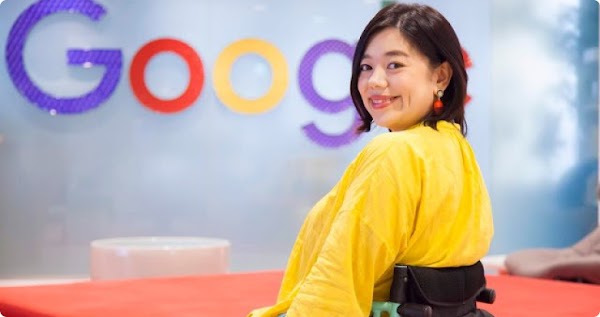 A woman with dark hair wearing a yellow shirt sits in a wheelchair and looks over her left shoulder at the camera. Behind her is a wall with the Google logo.